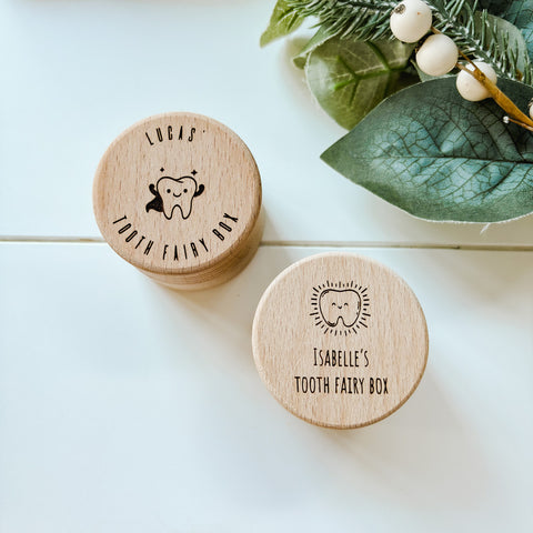 Two wooden tooth fairy boxes with personalized names on a light surface with greenery.